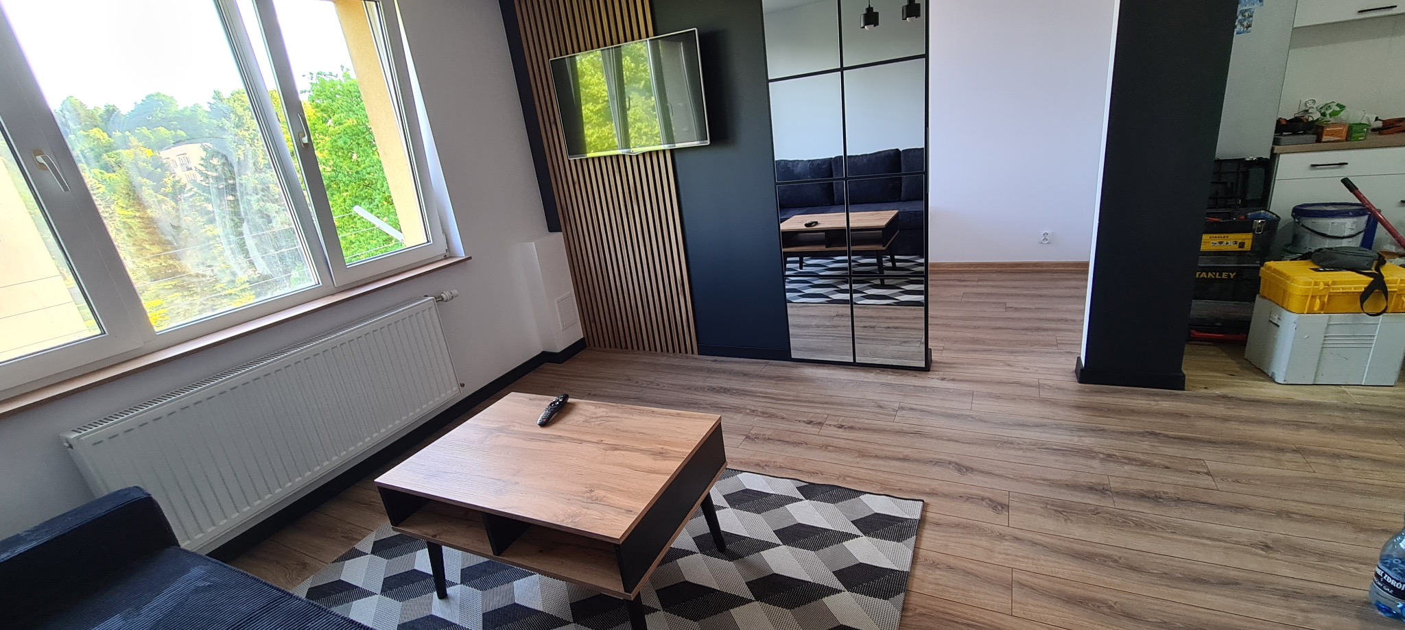 Nowoczesny salon po remoncie z drewnianą podłogą, ścianą z lamelami i lustrzanymi drzwiami przesuwnymi, odbijającymi wnętrze pokoju. Na pierwszym planie stolik kawowy z pilotem i geometryczny dywan.