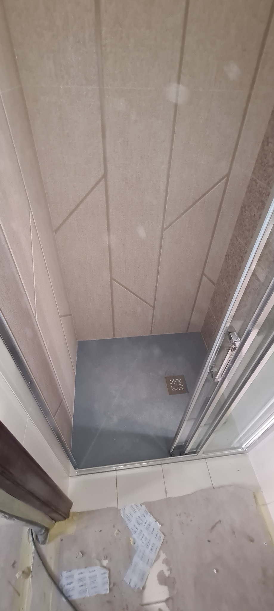 Wnętrze kabiny prysznicowej z szarym brodzikiem i beżowymi płytkami ściennymi w geometryczne wzory, widoczne pozostałości po pracach wykończeniowych na podłodze przed kabiną.
