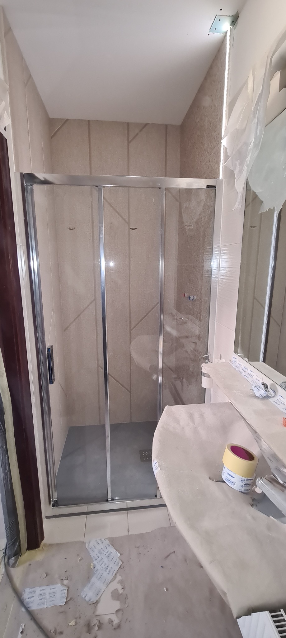 Nowoczesna kabina prysznicowa z przesuwnymi drzwiami w trakcie montażu, widoczne zabezpieczenia folią i taśma malarska na umywalce, jasne płytki z geometrycznym wzorem na ścianach.