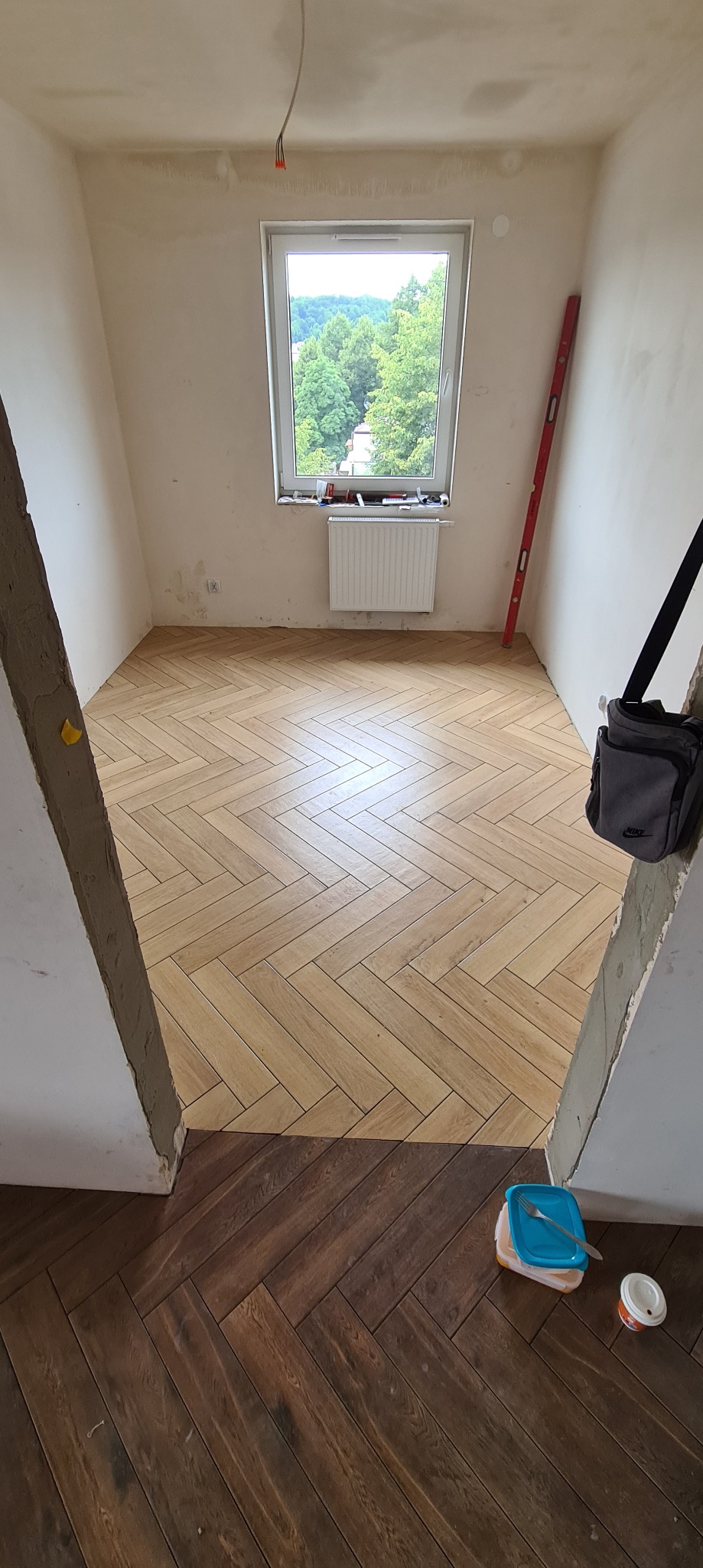 Jasna, drewnopodobna podłoga ułożona we wzór jodełki francuskiej w wyremontowanym pokoju z oknem, widoczne niedokończone ściany i poziomica oparta o ścianę, oraz pudełko z jedzeniem i torba robocza.