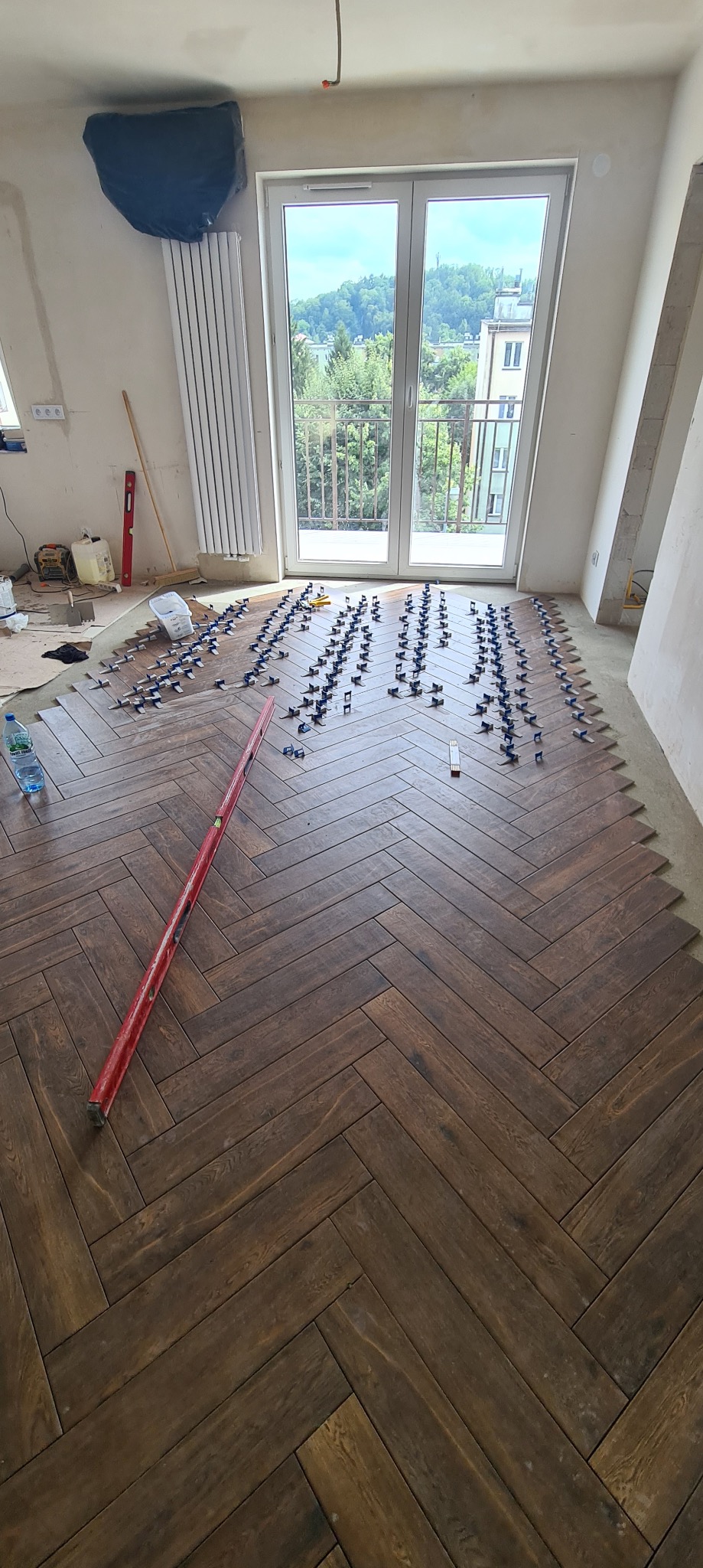 Układanie ciemnobrązowego parkietu w jodełkę francuską, widoczne kliny poziomujące, poziomica czerwona, okno balkonowe z widokiem na zieleń, prace w toku.