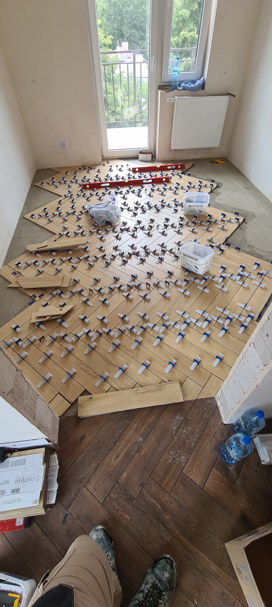 Układanie jasnych płytek podłogowych w jodełkę z użyciem systemu poziomowania, widoczne poziomice, pojemniki z akcesoriami i częściowo ułożona ciemna podłoga w jodełkę przy ścianie.