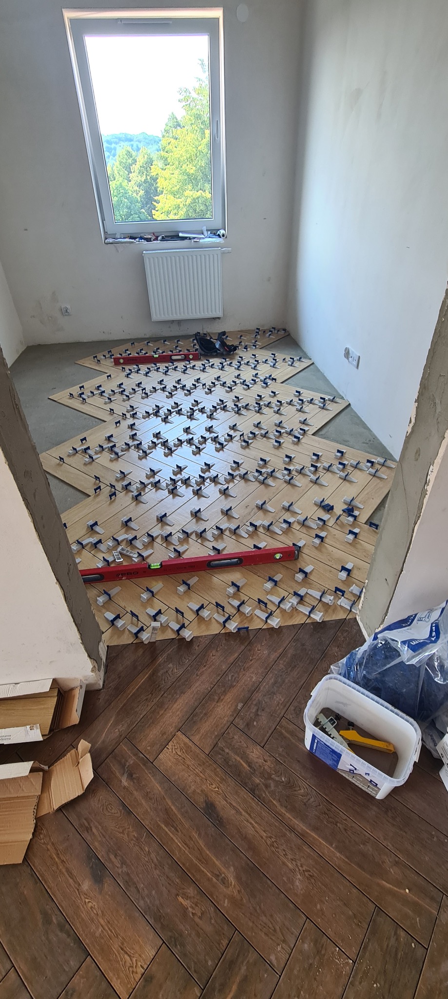 Układanie płytek imitujących parkiet w jodełkę; widoczne poziomica, system poziomowania płytek, oraz częściowo ułożona podłoga w drugim pomieszczeniu