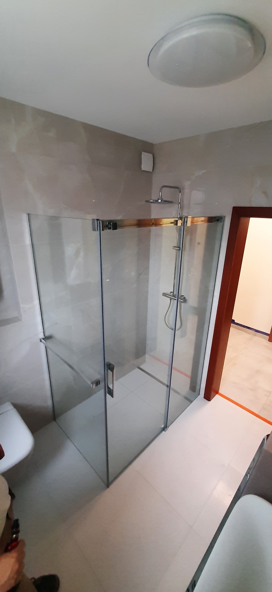 Szklana kabina prysznicowa z chromowanymi okuciami w jasnej łazience z marmurowymi płytkami, widoczny fragment toalety i drzwi do innego pomieszczenia z pomarańczowym zabezpieczeniem podłogi.