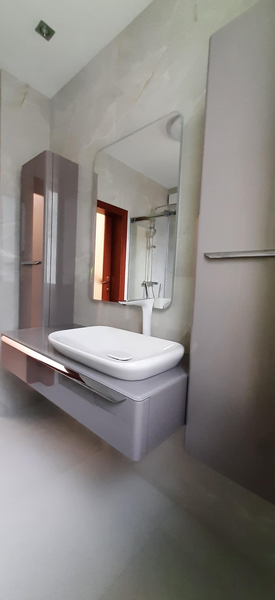 Nowoczesna łazienka z umywalką nablatową, szafką pod umywalką w kolorze taupe z miedzianymi akcentami, lustrem i wysoką szafką, widok na kabinę prysznicową w odbiciu lustra.