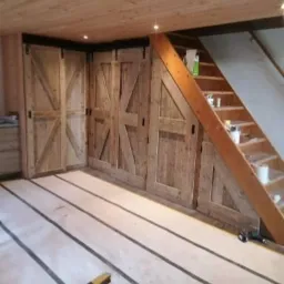 Drewniane, przesuwne drzwi do zabudowy pod skosem schodów, z dekoracyjnym wzorem w kształcie litery X, zamontowane na czarnych szynach, widoczne na tle surowego, drewnianego sufitu i przygotowanej...