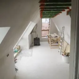 Poddasze w trakcie prac wykończeniowych, widoczne ocieplone krokwie, przygotowane ściany do malowania, piec wolnostojący i rozłożone narzędzia.