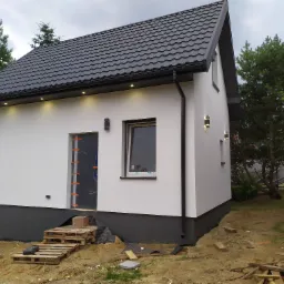 Nowoczesny, biały dom z ciemną, metalową dachówką i oświetleniem elewacyjnym, widoczne detale wykończenia fundamentów i przygotowane wejście.