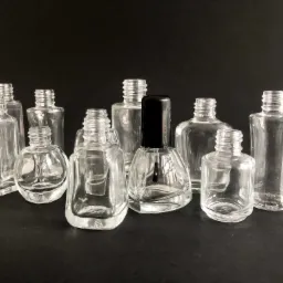 Grupa pustych, szklanych buteleczek o różnych kształtach, prawdopodobnie przeznaczonych na lakiery do paznokci, na czarnym tle. Jedna buteleczka ma czarny korek z pędzelkiem.