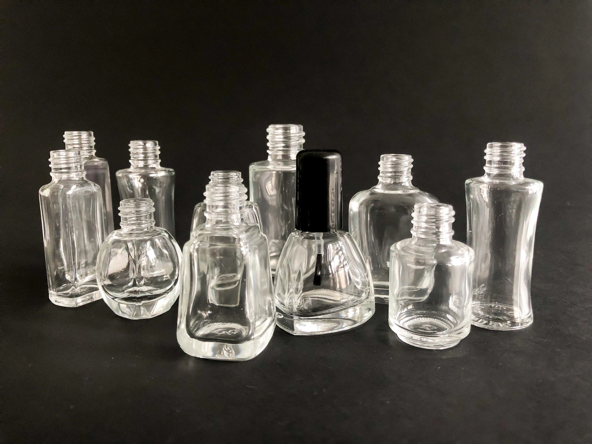 Grupa pustych, szklanych buteleczek o różnych kształtach, prawdopodobnie przeznaczonych na lakiery do paznokci, na czarnym tle. Jedna buteleczka ma czarny korek z pędzelkiem.