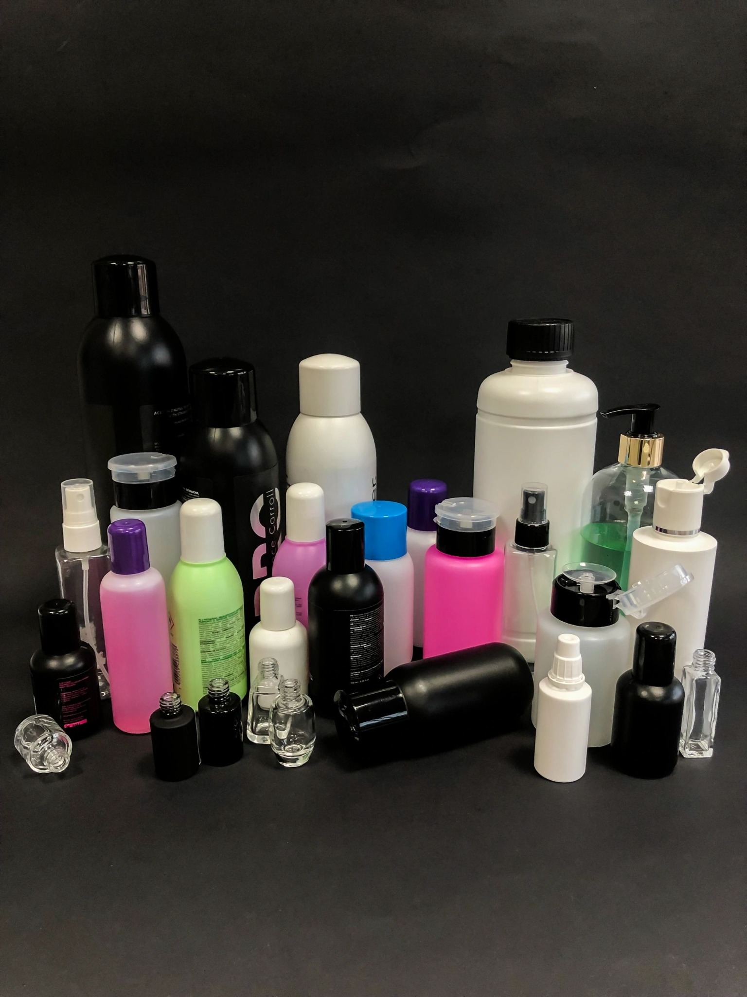 Różnorodne butelki i pojemniki kosmetyczne w kolorach czarnym, białym, różowym i zielonym, przeznaczone do pakowania produktów kosmetycznych w Ciechanowie, na jednolitym czarnym tle.