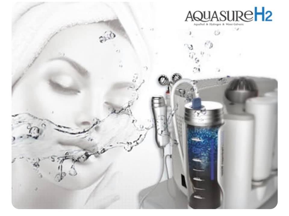 Kobieta z zamkniętymi oczami, na twarzy rozpryskująca się woda, obok urządzenie kosmetyczne Aquasure H2 z pojemnikiem wypełnionym niebieskimi elementami.