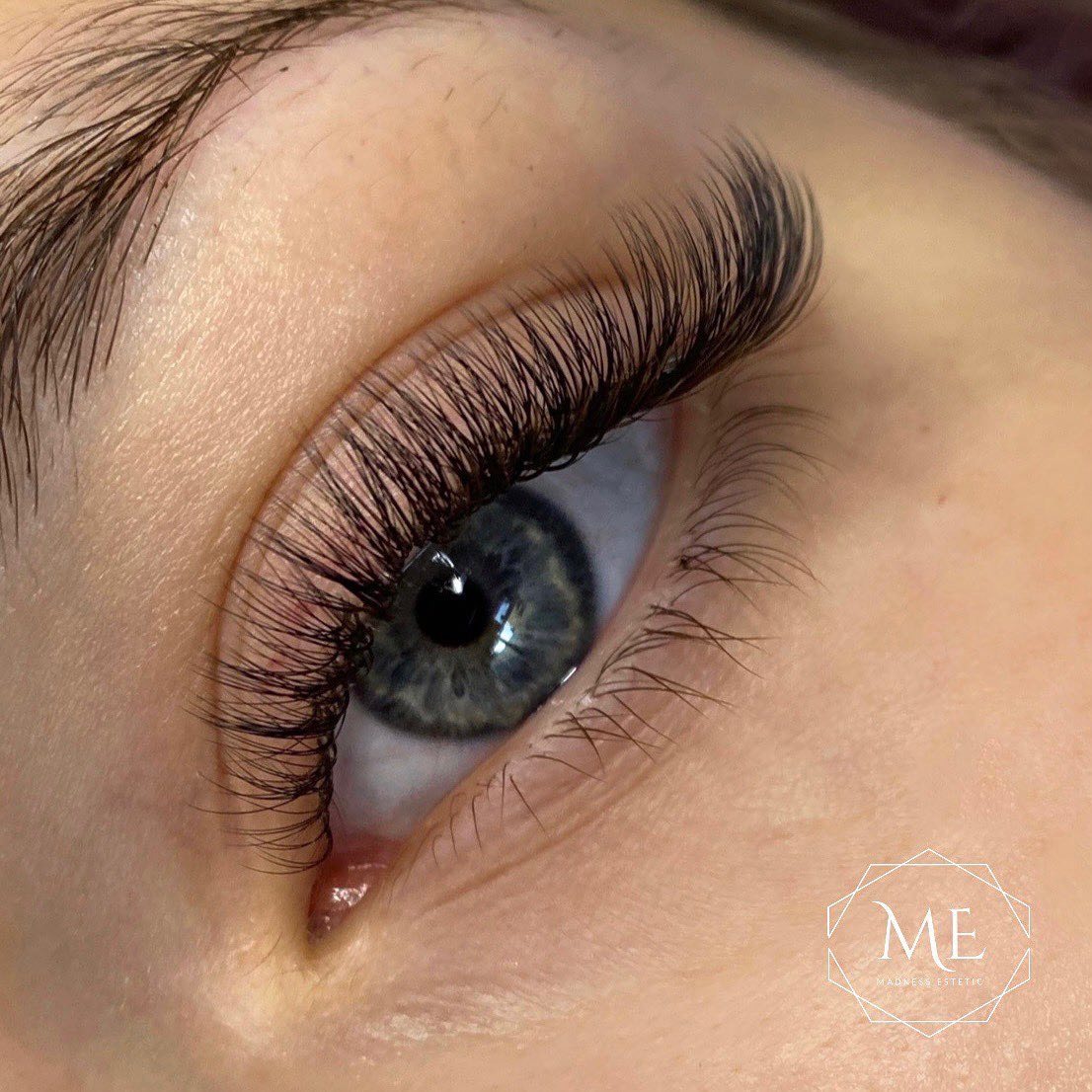 Zbliżenie na oko z długimi, gęstymi, profesjonalnie przedłużonymi rzęsami; widoczny subtelny makijaż i logo salonu kosmetycznego w prawym dolnym rogu.