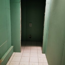 Technics Group - Wnętrze łazienki w trakcie remontu, ściany wyłożone zieloną płytą gipsowo-kartonową, widoczne łączenia i ślady po wkrętach, na podłodze białe, kwadratowe płytki ceramiczne, w głębi otwór...