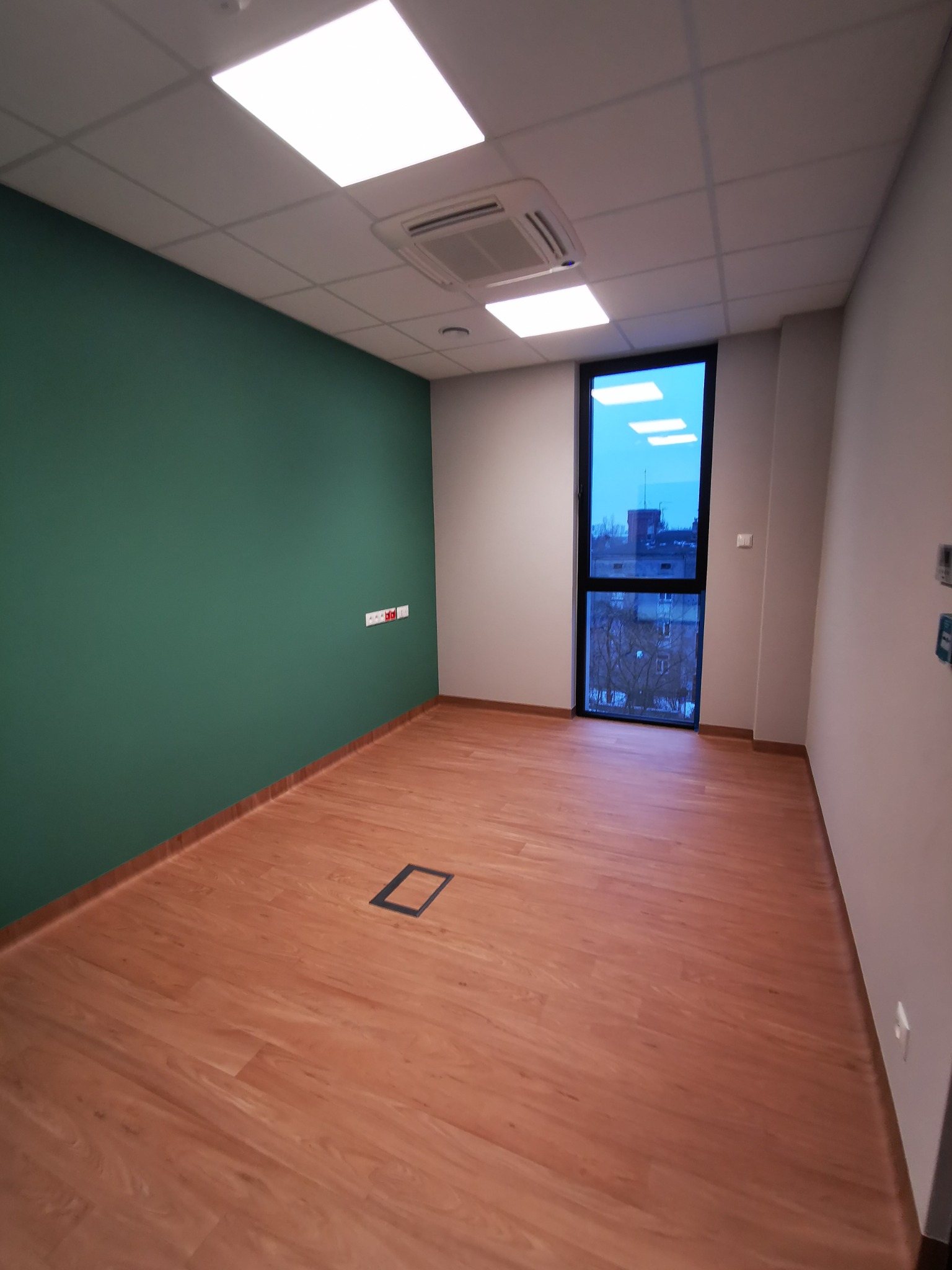 Puste pomieszczenie z panelami podłogowymi, pomalowaną na zielono ścianą i oknem z widokiem na miasto. Widoczne gniazdka elektryczne i kratka wentylacyjna w podłodze.