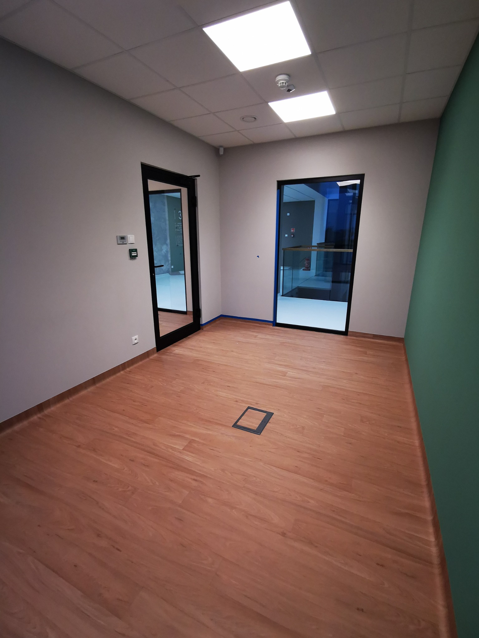 Wnętrze biurowe po remoncie z panelami podłogowymi o wzorze drewna, szarą ścianą z włącznikiem i gniazdkiem, zieloną ścianą, dwoma wejściami z czarnymi futrynami oraz kwadratową puszką podłogową.