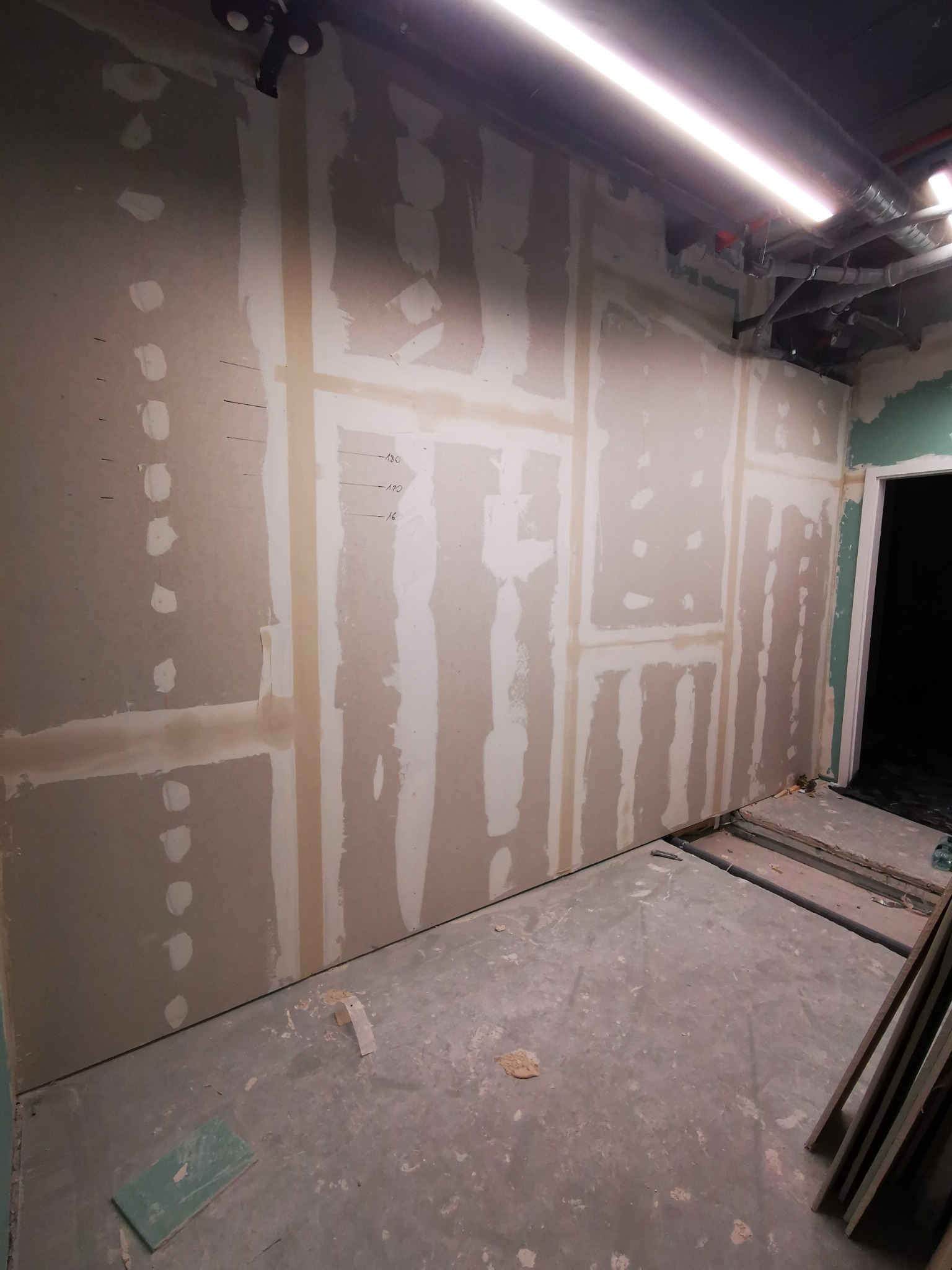 Montaż płyt gipsowo-kartonowych na ścianie z widocznymi łączeniami i wkrętami, przygotowanymi do szpachlowania, na podłodze widoczne resztki materiałów i narzędzia.