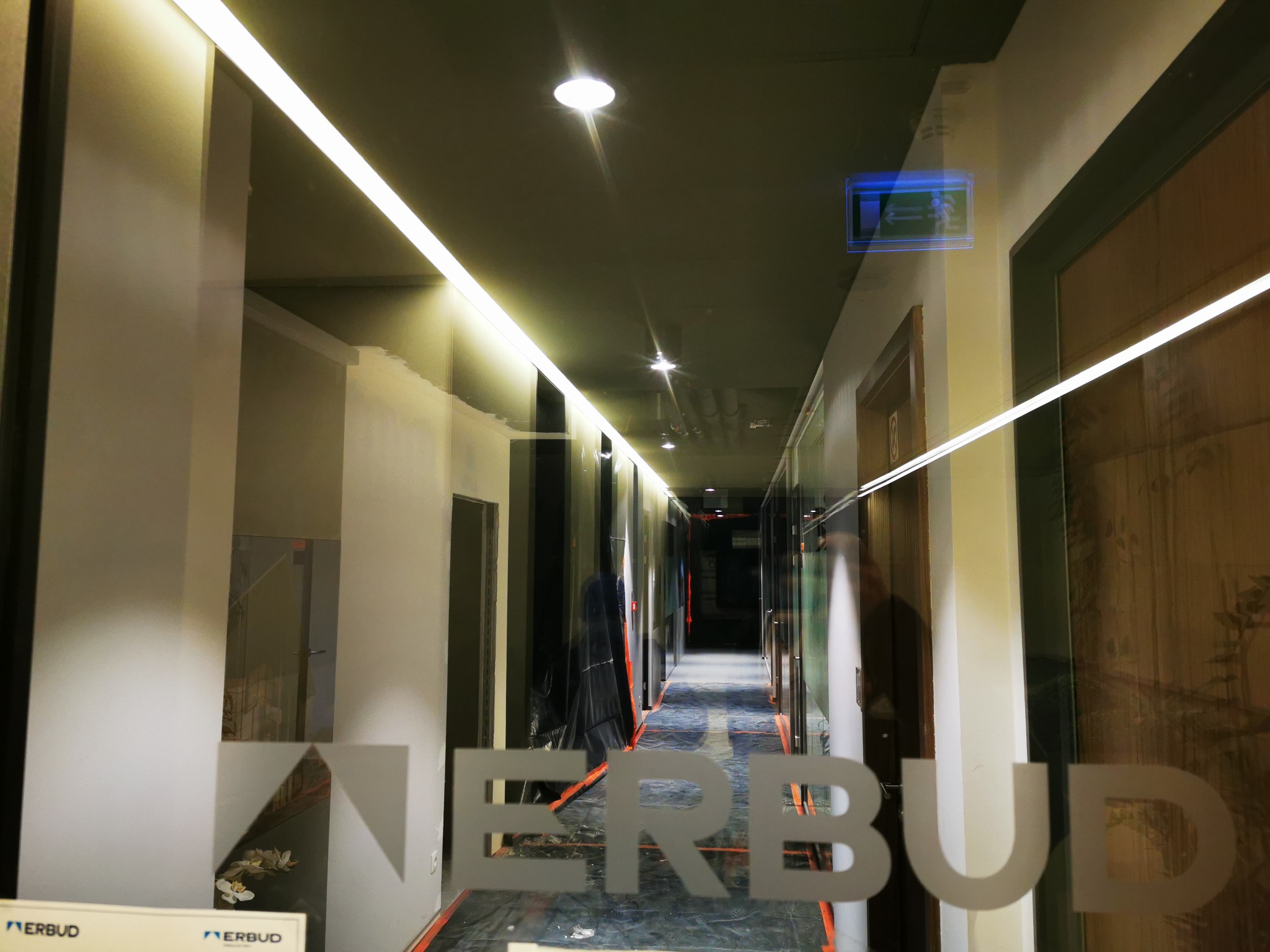 Wnętrze korytarza w trakcie remontu, widoczne zabezpieczenia podłogi folią, logo firmy Erbud na szybie, oświetlenie ledowe sufitowe i punktowe.