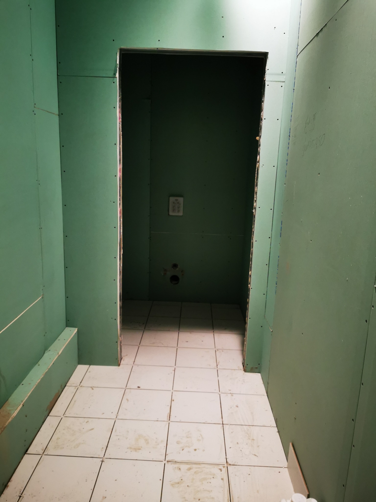 Wnętrze łazienki w trakcie remontu, ściany wyłożone zieloną płytą gipsowo-kartonową, widoczne łączenia i ślady po wkrętach, na podłodze białe, kwadratowe płytki ceramiczne, w głębi otwór...