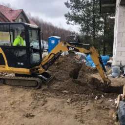 Żółto-czarna mini koparka CAT 301.8C wykopuje rów w ziemi na placu budowy domu jednorodzinnego; operator w jaskrawej kurtce, w tle przenośna toaleta, materiały budowlane i drzewa.