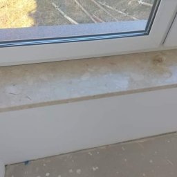 Ideal Rafał Dyro - Jasny parapet wewnętrzny o marmurkowej fakturze, zamontowany w białym oknie PCV, z widocznymi śladami prac wykończeniowych na podłodze.
