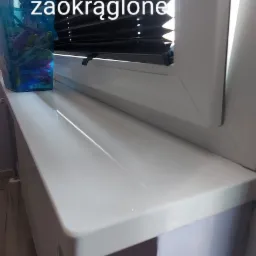 Biały, błyszczący parapet z zaokrąglonym brzegiem, zamontowany w oknie z żaluzjami plisowanymi. Na parapecie widoczny pojemnik z klamerkami do bielizny.