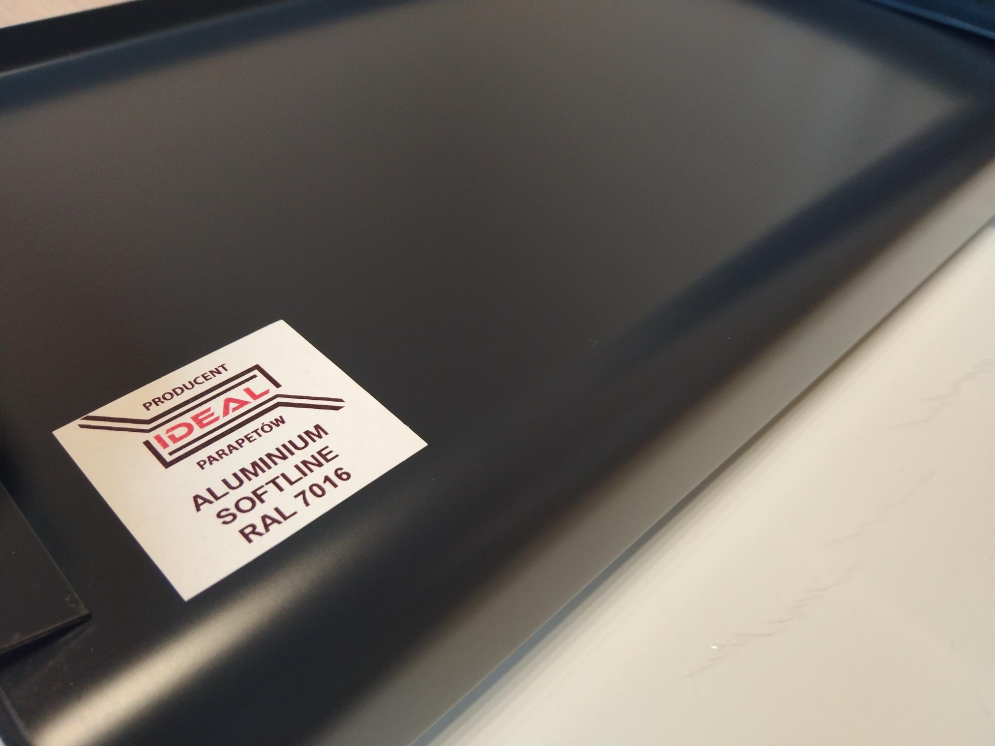 Szary aluminiowy parapet z naklejką producenta 'Ideal Parapetów Aluminium Softline RAL 7016' na białym blacie.