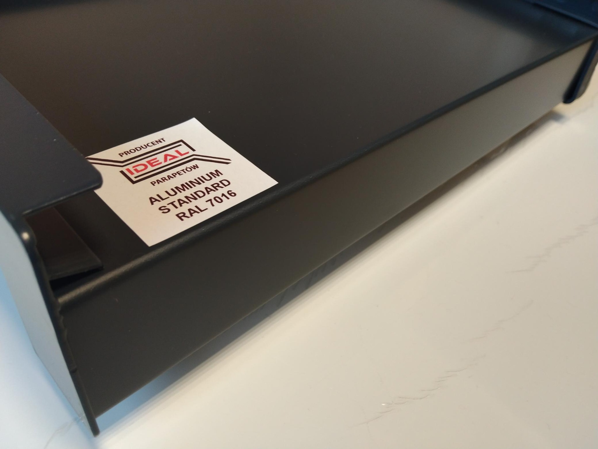 Fragment czarnego, aluminiowego parapetu z naklejoną etykietą producenta 'IDEAL' z informacją 'Aluminium Standard RAL 7016'.