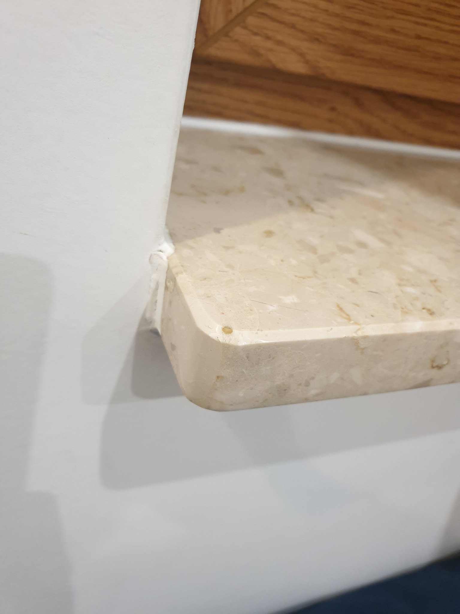 Beżowy parapet z widoczną strukturą kamienia, zamontowany przy białej ścianie i drewnianym elemencie, uszczelniony silikonem.