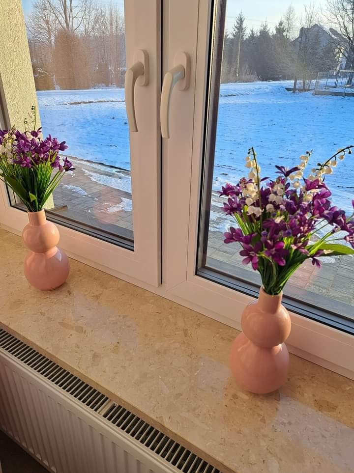 Beżowy parapet z widocznym wzorem, na którym stoją dwie różowe wazy z fioletowymi kwiatami, za oknem zimowy krajobraz.
