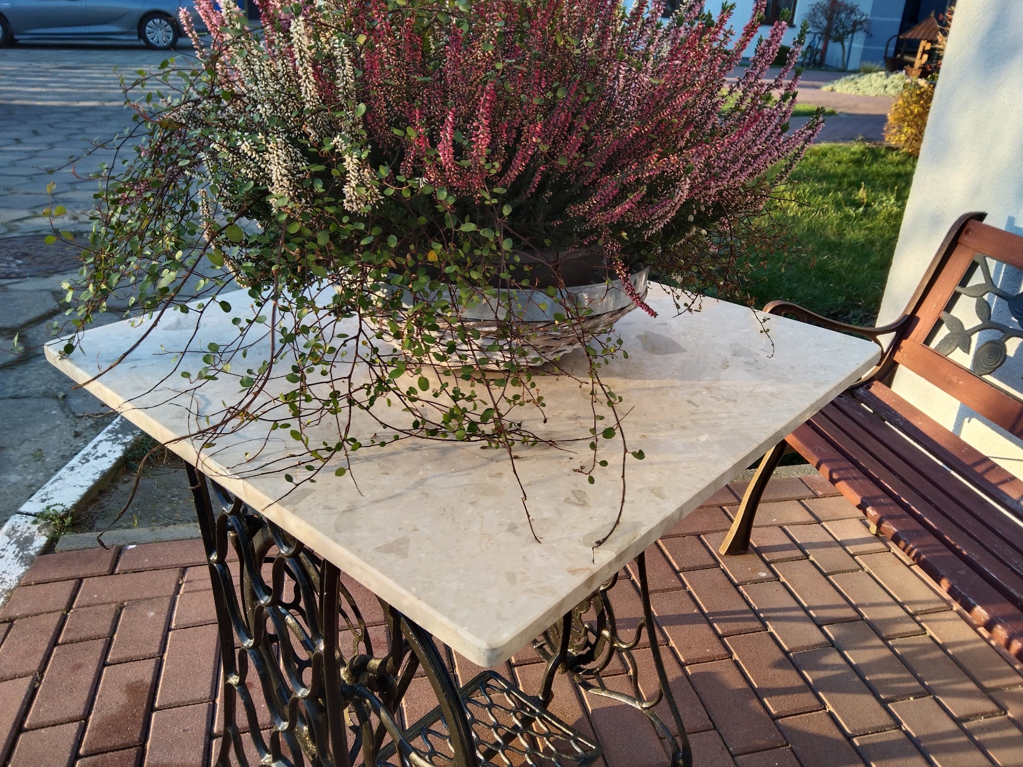 Kwadratowy blat z jasnego konglomeratu na ozdobnej, metalowej podstawie maszyny do szycia, używany jako stolik na zewnątrz, z doniczką wrzosów i pnączy na wierzchu, obok drewnianej ławki.
