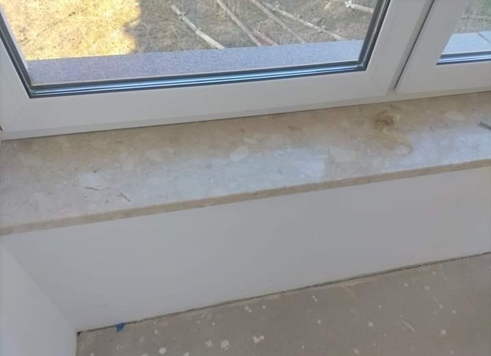 Jasny parapet wewnętrzny o marmurkowej fakturze, zamontowany w białym oknie PCV, z widocznymi śladami prac wykończeniowych na podłodze.