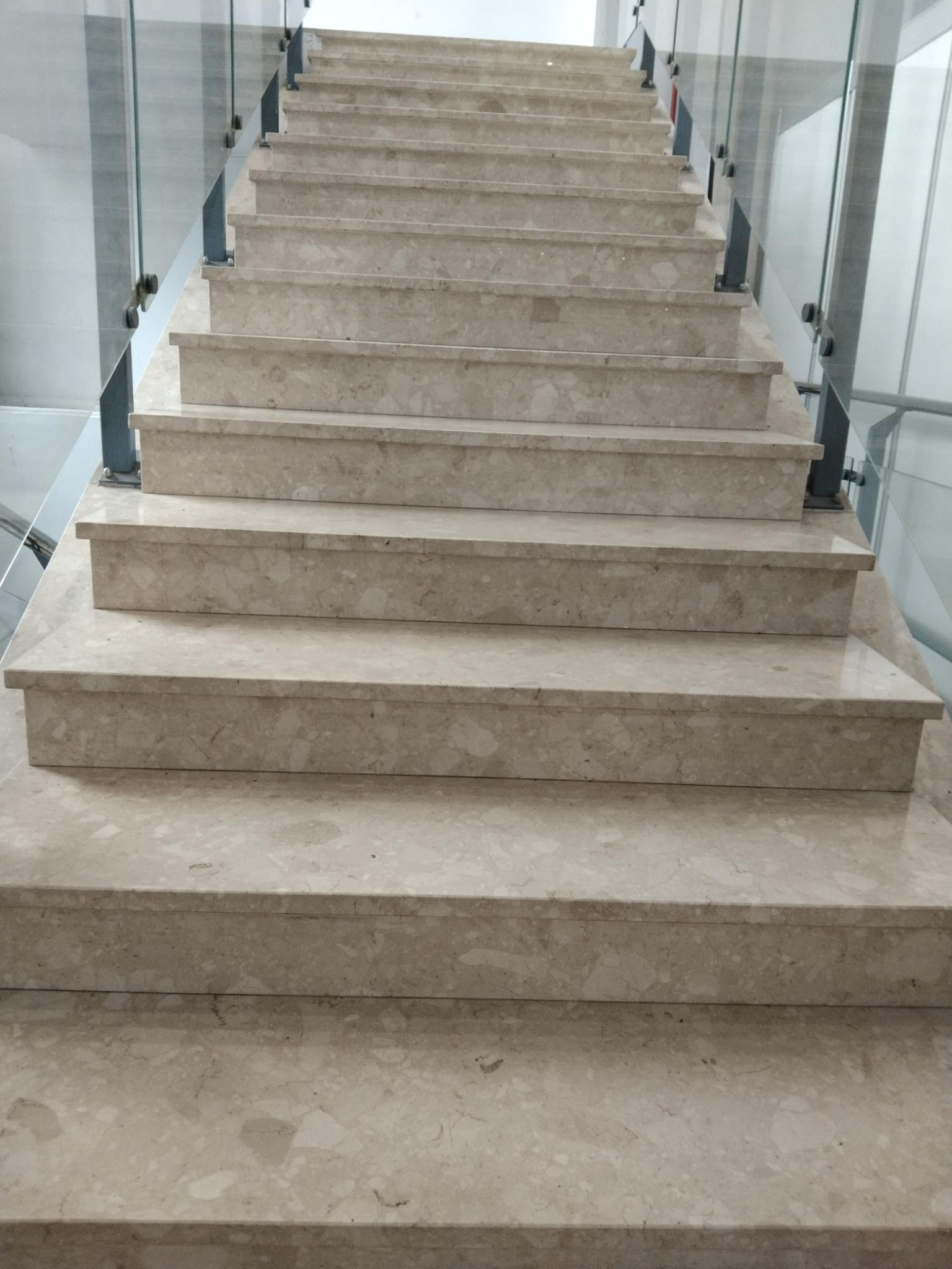 Schody wewnętrzne wyłożone jasnym konglomeratem z delikatnym wzorem, zabezpieczone szklaną balustradą mocowaną punktowo do stopni.