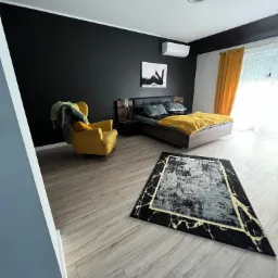 Nowoczesna sypialnia z czarną ścianą, drewnianym łóżkiem z żółtą narzutą, żółtym fotelem i dywanem w stylu marmuru na podłodze z paneli imitujących drewno.