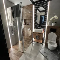 Nowoczesna łazienka z kabiną prysznicową typu walk-in, drewnopodobnymi płytkami w strefie prysznica i pod umywalką, szarą armaturą, podświetleniem LED i dekoracyjną ścianą z wzorem szachownicy za...