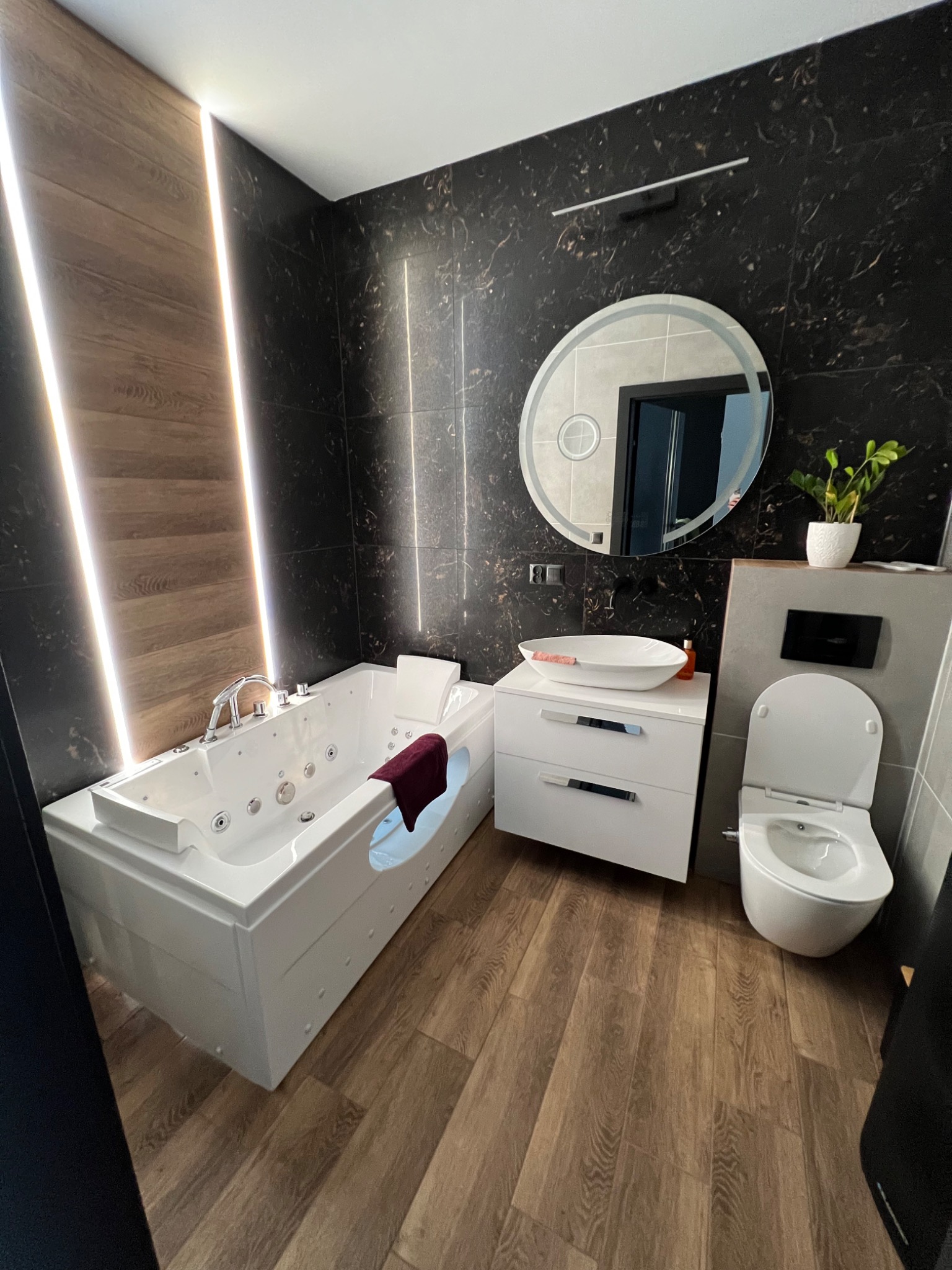 Nowoczesna łazienka z wanną jacuzzi, okrągłym lustrem z podświetleniem LED, umywalką nablatową na szafce z szufladami, toaletą podwieszaną i drewnopodobną podłogą, oświetlona pionowymi paskami LED.