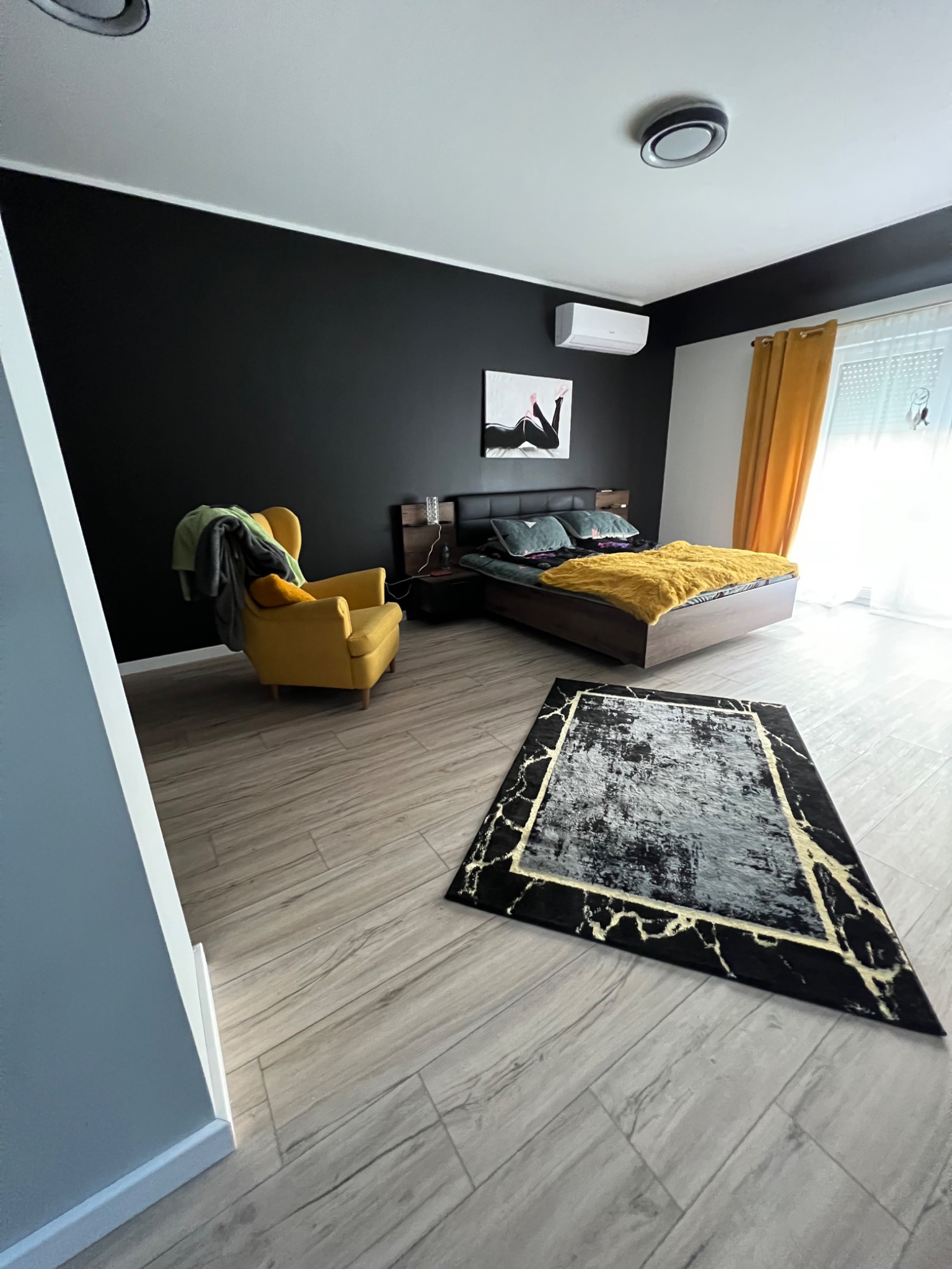 Nowoczesna sypialnia z czarną ścianą, drewnianym łóżkiem z żółtą narzutą, żółtym fotelem i dywanem w stylu marmuru na podłodze z paneli imitujących drewno.