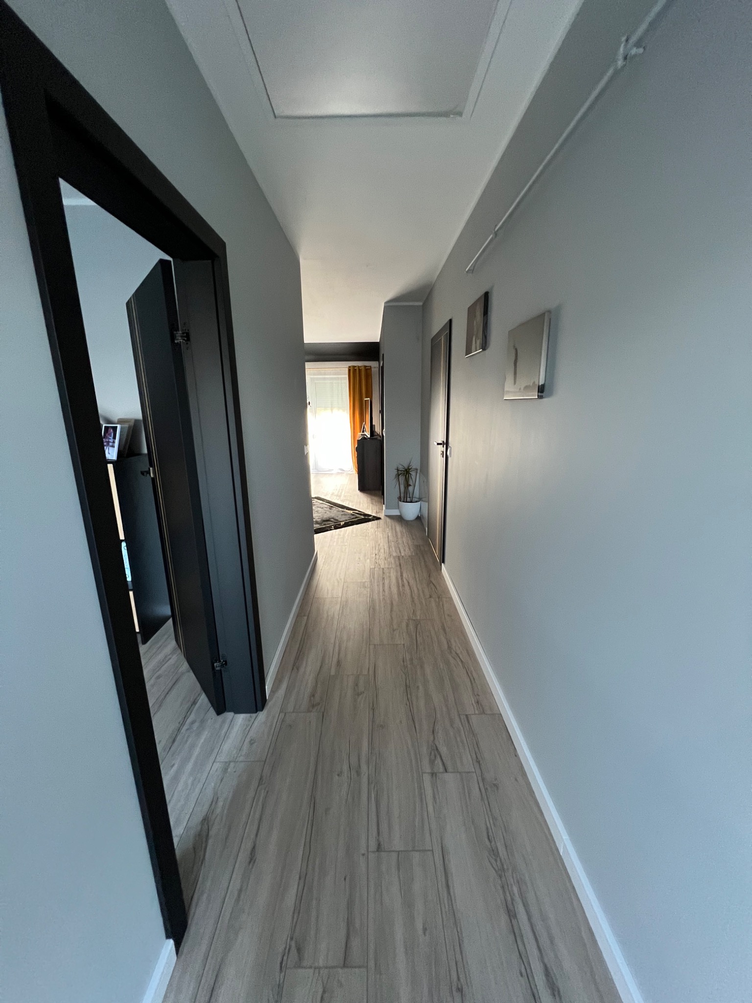 Długi korytarz z szarą podłogą imitującą drewno, szare ściany z trzema obrazami, czarne ramy drzwi i widok na pokój z żółtymi zasłonami.