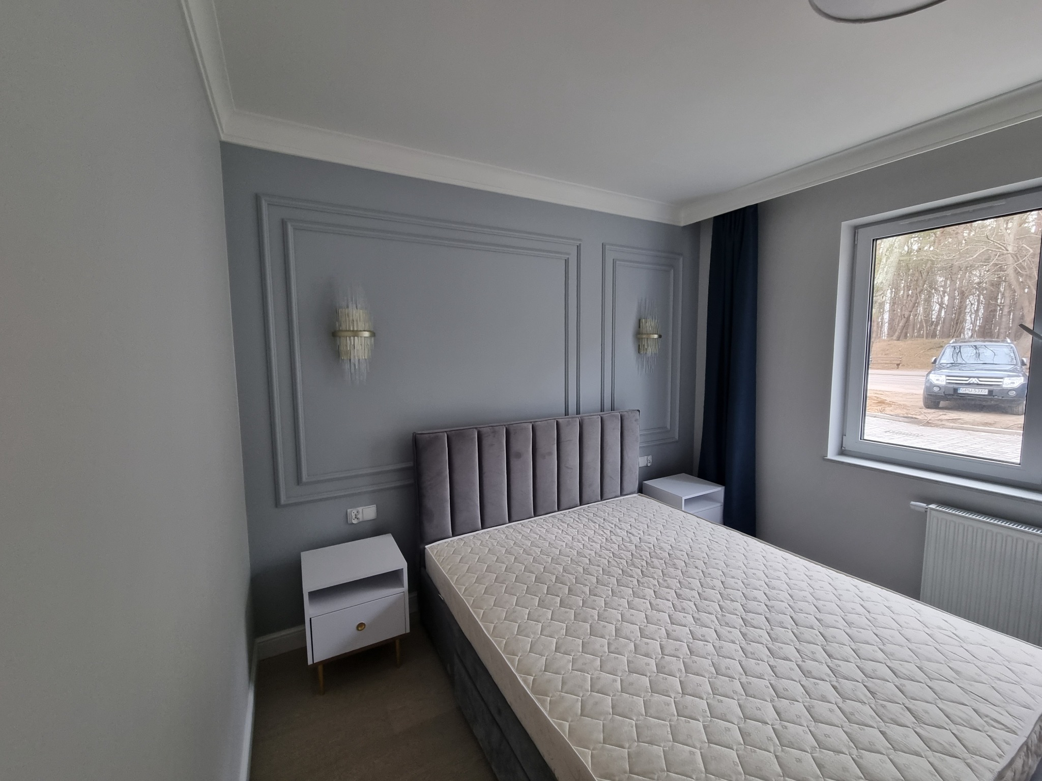 Sypialnia w odcieniach szarości z łóżkiem kontynentalnym, dwiema szafkami nocnymi i dekoracyjnymi panelami ściennymi z oświetleniem. Za oknem widoczny samochód.