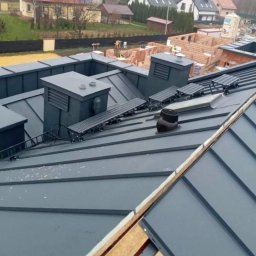 E&D Complex - Szary dach z blachy na rąbek stojący z kominami, wyłazami dachowymi i zabezpieczeniami przeciwśniegowymi, widok z góry na tle budynków w budowie.