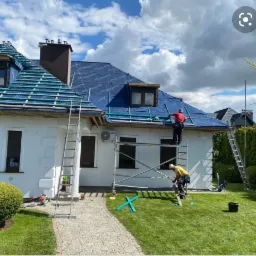 Dwóch dekarzy na rusztowaniu i drabinie montuje folię na dachu białego domu z dwoma lukarnami, widoczne przygotowane łaty, trawnik i kostka brukowa.