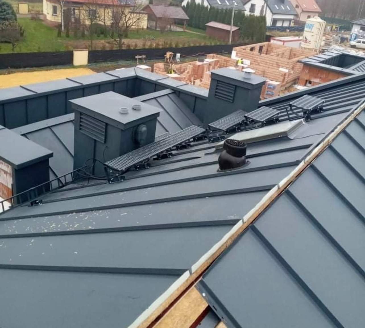 Szary dach z blachy na rąbek stojący z kominami, wyłazami dachowymi i zabezpieczeniami przeciwśniegowymi, widok z góry na tle budynków w budowie.