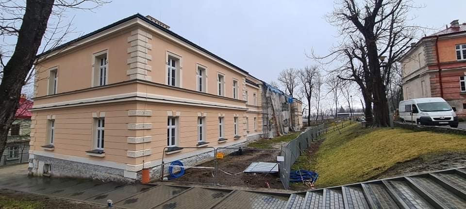 Budynek po renowacji elewacji w odcieniach beżu z białymi detalami architektonicznymi, widoczne rusztowania i zabezpieczenia na części budynku, sugerujące trwające prace.