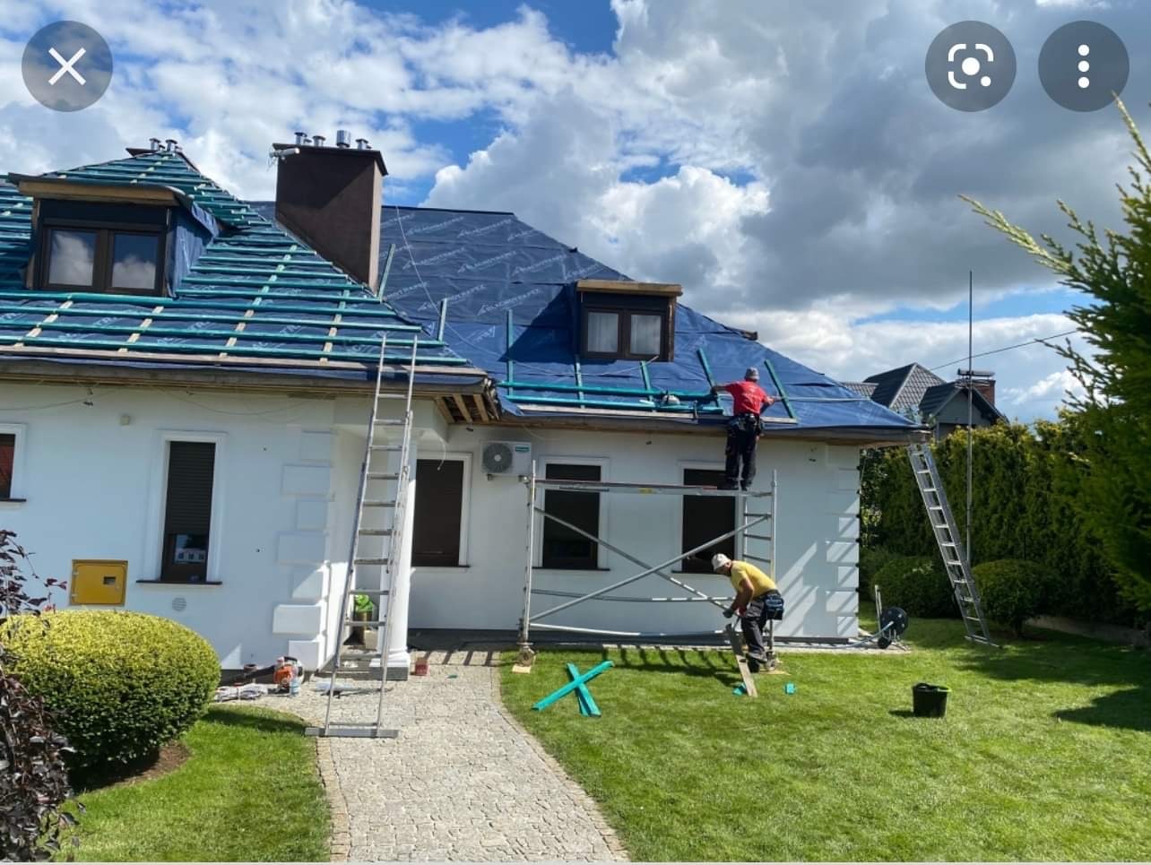 Dwóch dekarzy na rusztowaniu i drabinie montuje folię na dachu białego domu z dwoma lukarnami, widoczne przygotowane łaty, trawnik i kostka brukowa.