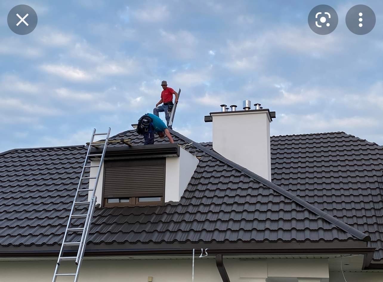 Dwóch mężczyzn na dachu domu z ciemną dachówką, jeden na drabinie przy kominie, drugi pochylony nad oknem dachowym z roletą zewnętrzną, widoczne narzędzia i drabiny.