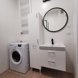 Zrobimy Firma Budowlana - Biała łazienka z pralką Beko, szafką umywalkową z czarną armaturą, okrągłym lustrem w czarnej ramie i grzejnikiem drabinkowym na ścianie z białych płytek.
