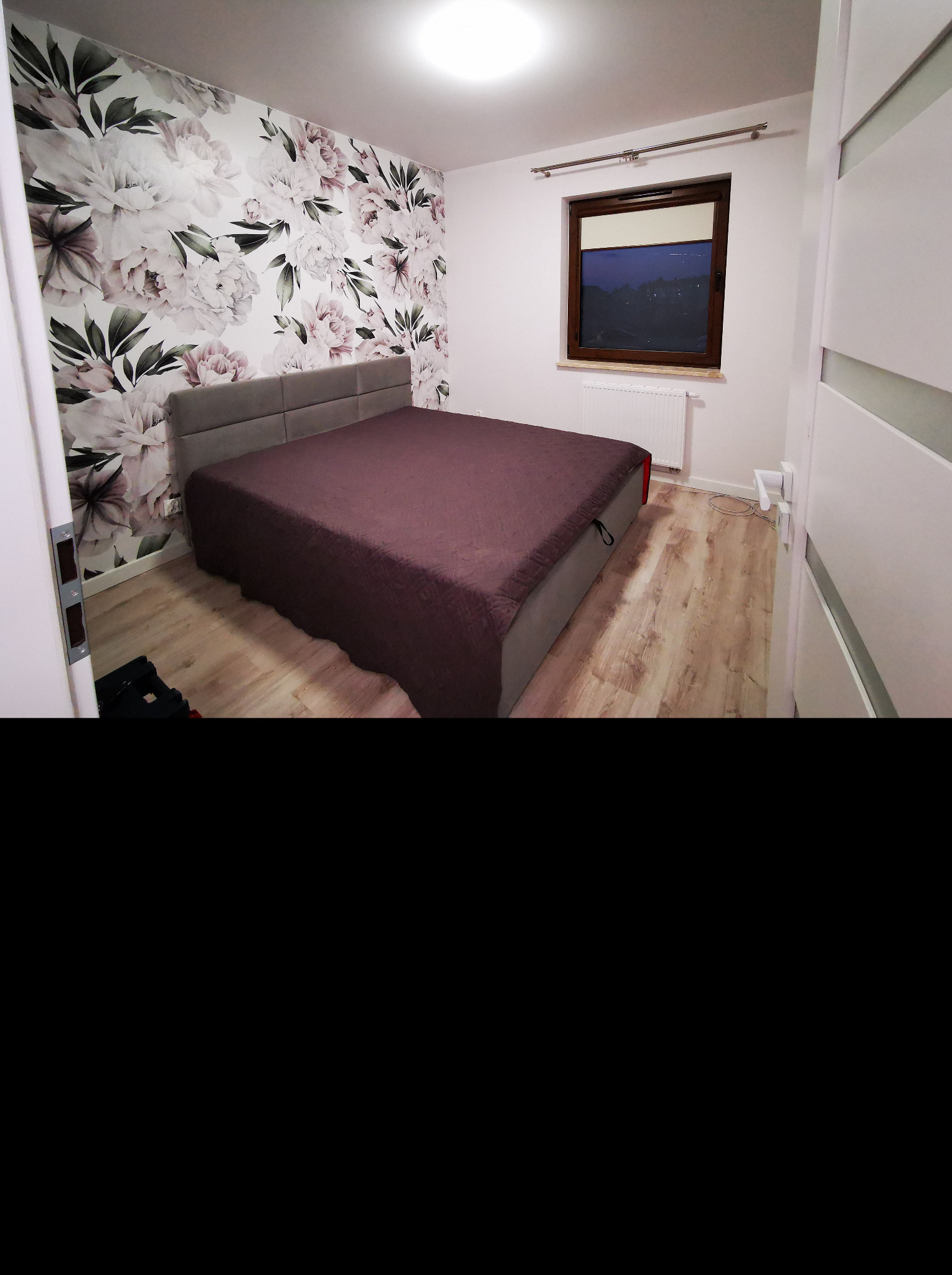 Sypialnia po remoncie: łóżko z szarym zagłówkiem i bordową narzutą, ściana w kwiatowe wzory, okno z drewnianą ramą i biały grzejnik.