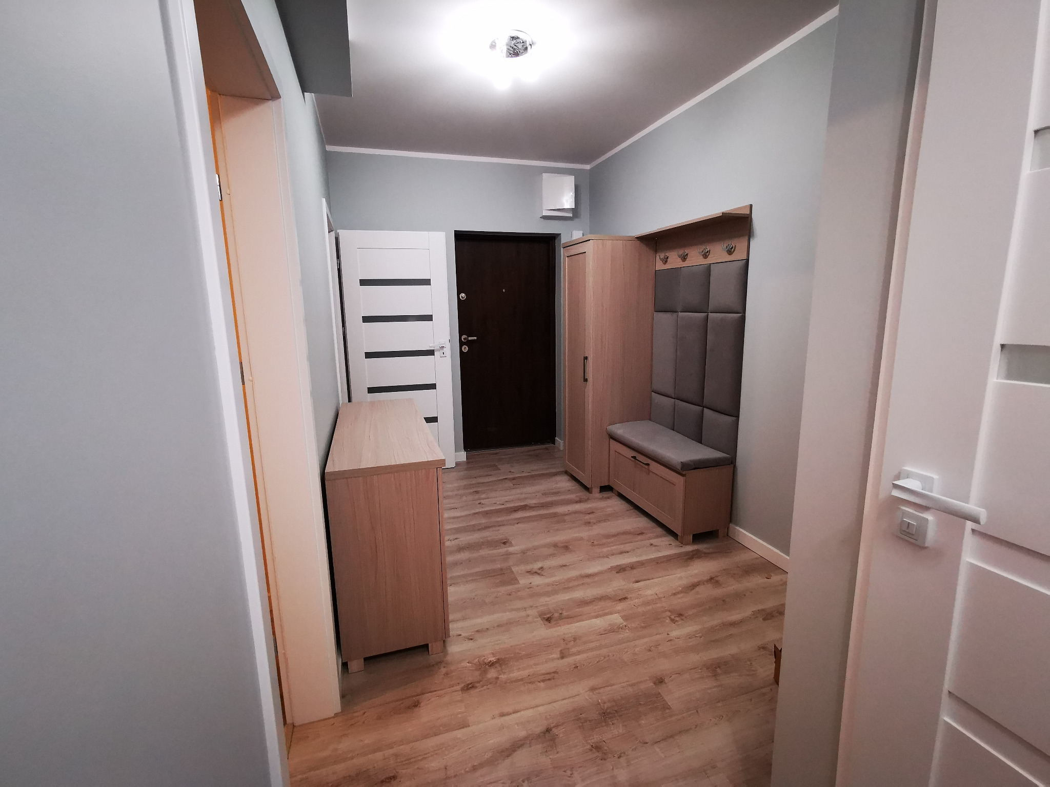 Przedpokój w odcieniach szarości i beżu z drewnianą podłogą, szafką na buty z siedziskiem i komodą z forniru drewnopodobnego, widok na drzwi wejściowe i wewnętrzne.