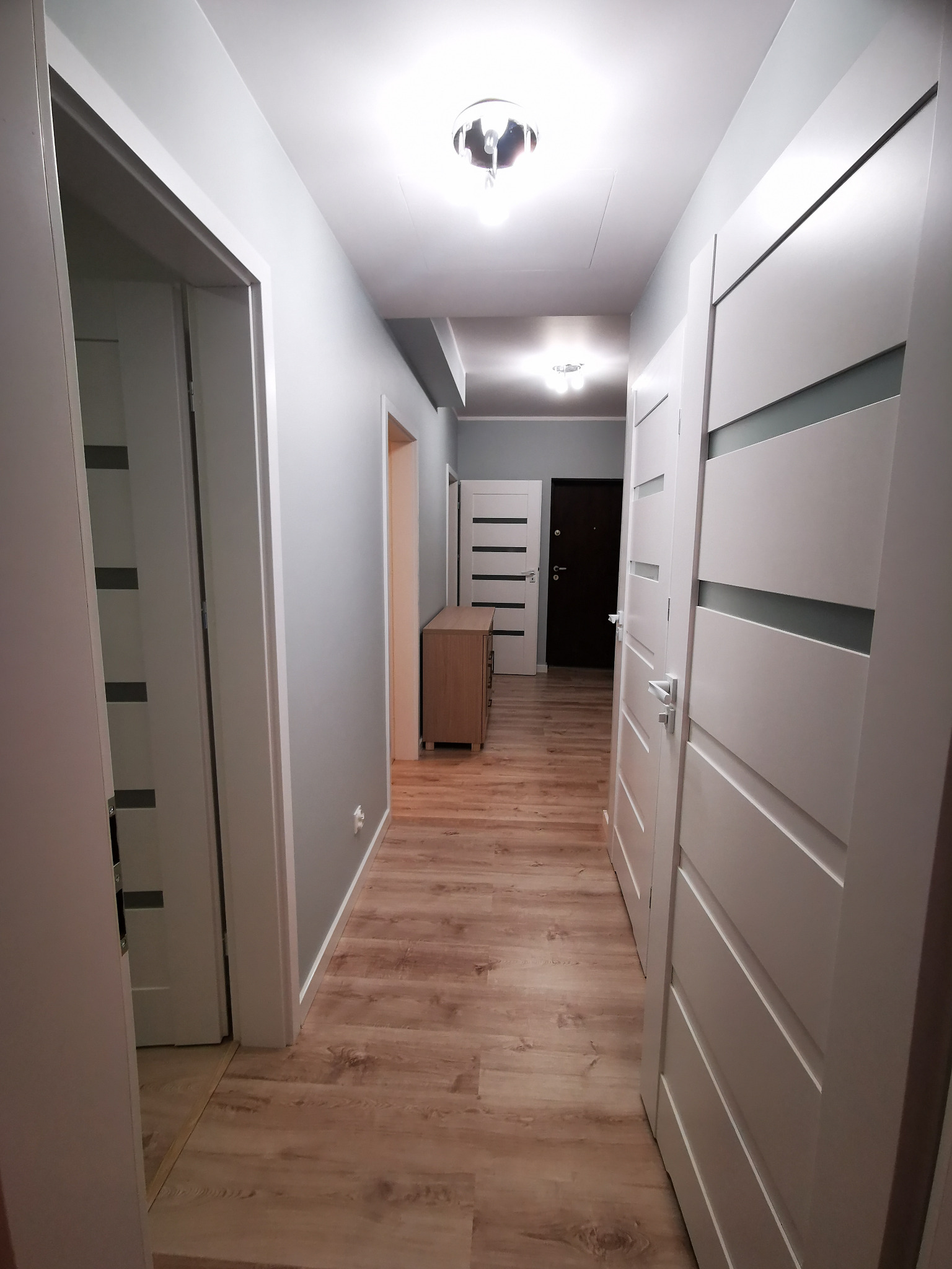 Wnętrze korytarza po remoncie z drewnopodobną podłogą, szarymi ścianami i białymi drzwiami z poziomymi przeszkleniami, oświetlonego dwoma lampami sufitowymi.