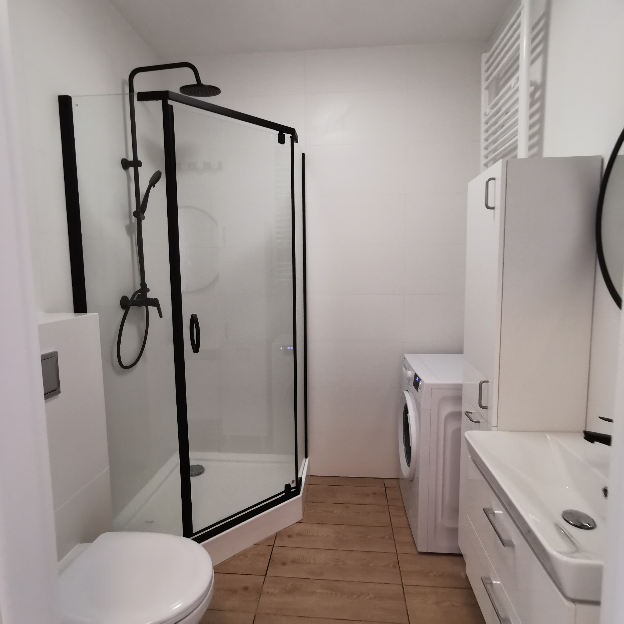 Nowoczesna łazienka w bieli z czarnymi akcentami: kabina prysznicowa typu walk-in z czarną ramą, toaleta, umywalka z szafką, pralka i grzejnik drabinkowy na tle białych płytek i drewnopodobnej...